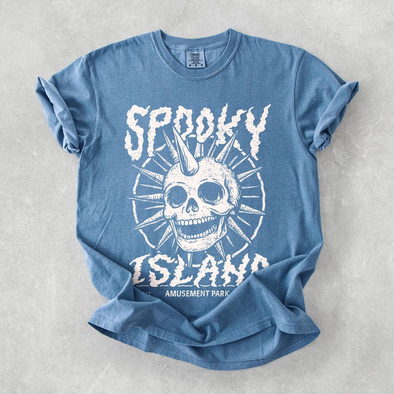 May include: A blue t-shirt with a white graphic featuring a skull with a horn, surrounded by spiky lines. The text "Spooky Island Amusement Park" is printed below the skull in a distressed font.