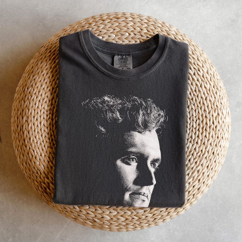 May include: Dark gray t-shirt featuring a black and white portrait of a man's face. The t-shirt is folded and placed on a round, woven, beige surface. The shirt has a crew neck and a tag at the neckline.