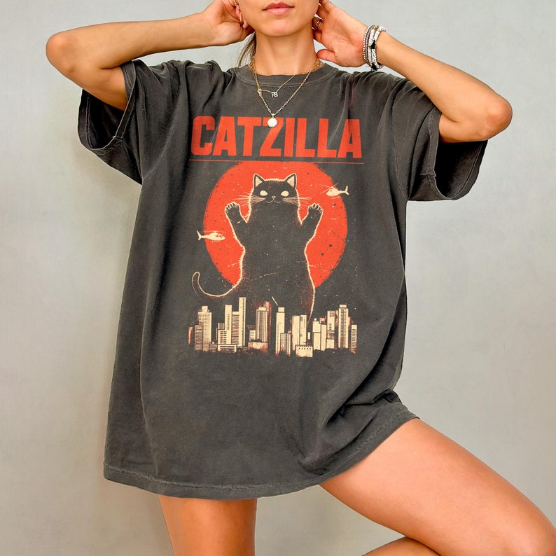 May include: A black oversized t-shirt with a graphic print of a black cat standing on a city skyline. The text "CATZILLA" is printed in white above the cat.