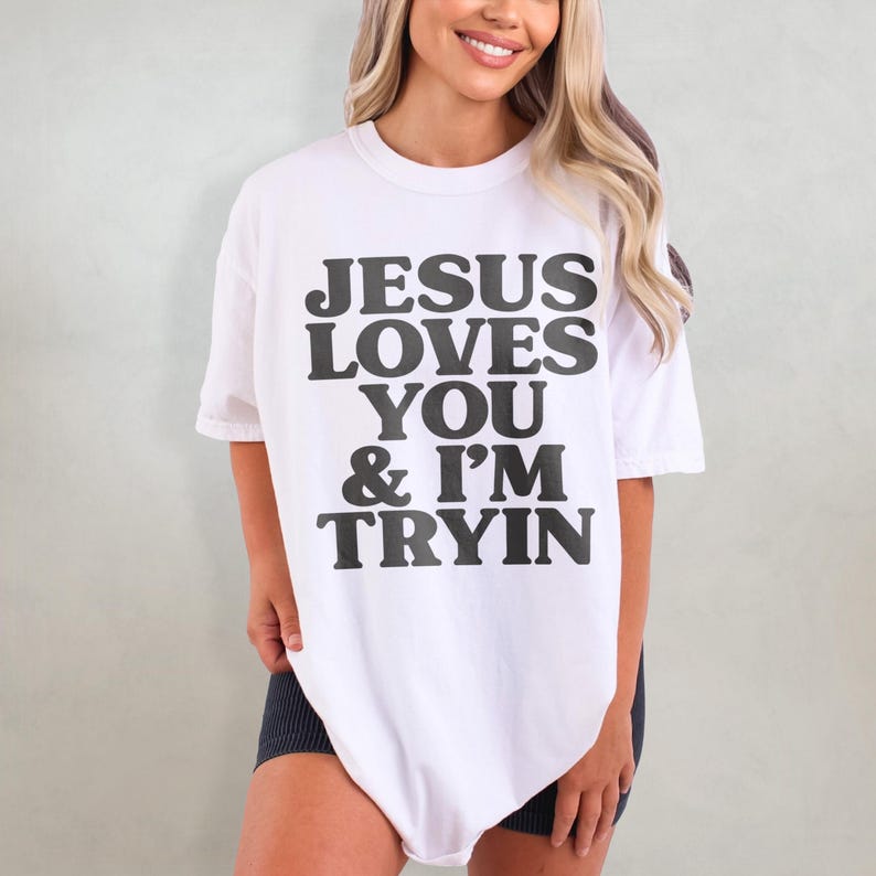 May include: White oversized t-shirt with black text that reads "JESUS LOVES YOU & I'M TRYIN"