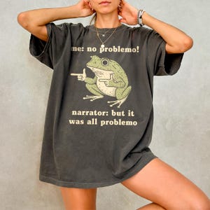 May include: Dark gray t-shirt featuring a green frog illustration with the text "me: no problemo!" and "narrator: but it was all problemo". The frog is pointing with one hand and giving a thumbs up with the other.