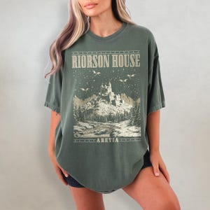 以下が含まれることがあります： 緑色のオーバーサイズTシャツ。雪景色の中に城が描かれたグラフィックプリントが施され、前面には「RIORSON HOUSE」と「ARETIA」の文字がプリントされています。