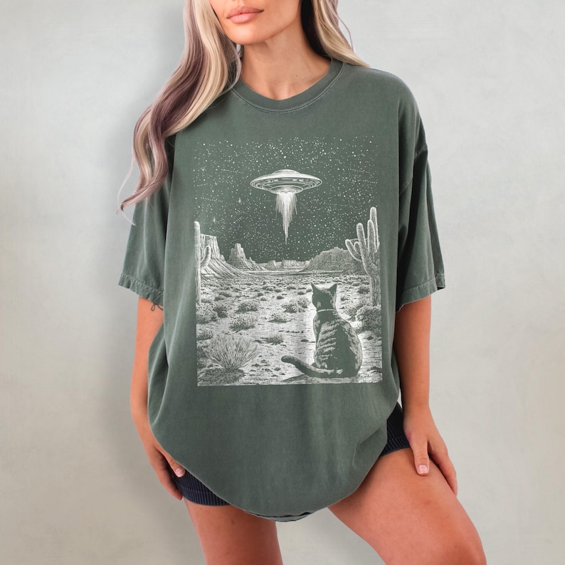 May include: A green t-shirt with a graphic print of a cat sitting in a desert landscape with a UFO in the sky. The graphic is in black and white.