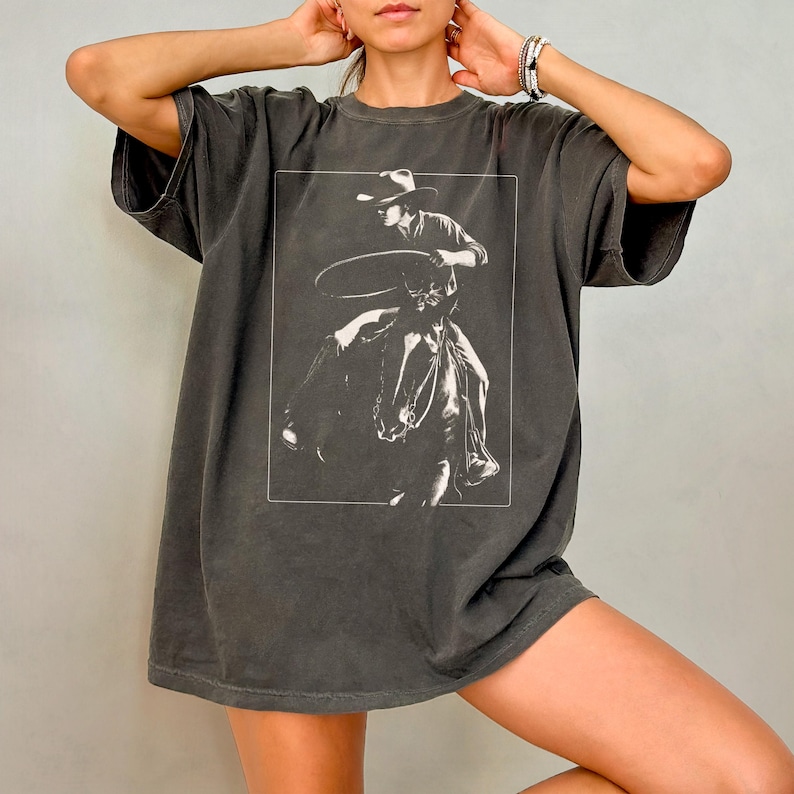 May include: A dark gray oversized t-shirt with a white graphic of a cowboy riding a horse and holding a lasso.