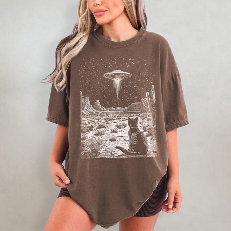 May include: A brown t-shirt with a graphic print of a cat sitting in a desert landscape with a UFO in the sky. The graphic is in white and black.
