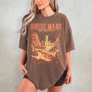 May include: Brown t-shirt with the text "HOUSE MARS THE INSTITUTE" in red and a graphic of a castle in a desert landscape. The shirt has short sleeves and a crew neck.