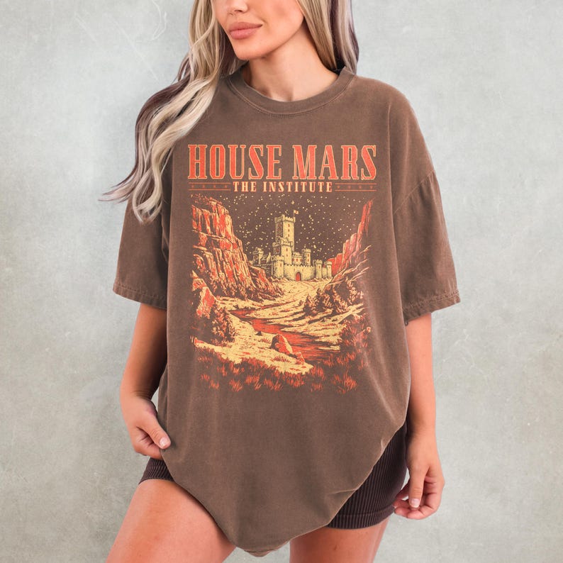 May include: Brown t-shirt with the text "HOUSE MARS THE INSTITUTE" in red and a graphic of a castle in a desert landscape. The shirt has short sleeves and a crew neck.