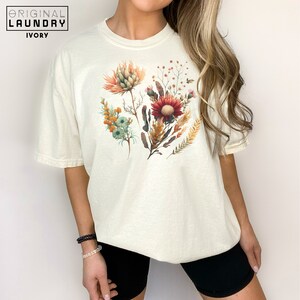 May include: A white t-shirt with a floral print featuring a variety of flowers in shades of red, orange, yellow, and green.