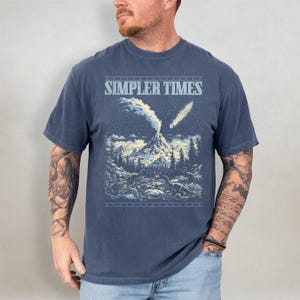 K&ouml;nnte beinhalten: Marineblaues T-Shirt mit dem Text "SIMPLER TIMES" &uuml;ber einer Berglandschaft mit Vulkan, B&auml;umen und Kometen. Die Grafik ist in Blau- und Wei&szlig;t&ouml;nen gehalten.