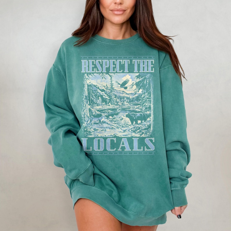 May include: A teal green sweatshirt with a vintage-style graphic print of a mountain scene and the text "Respect the Locals".