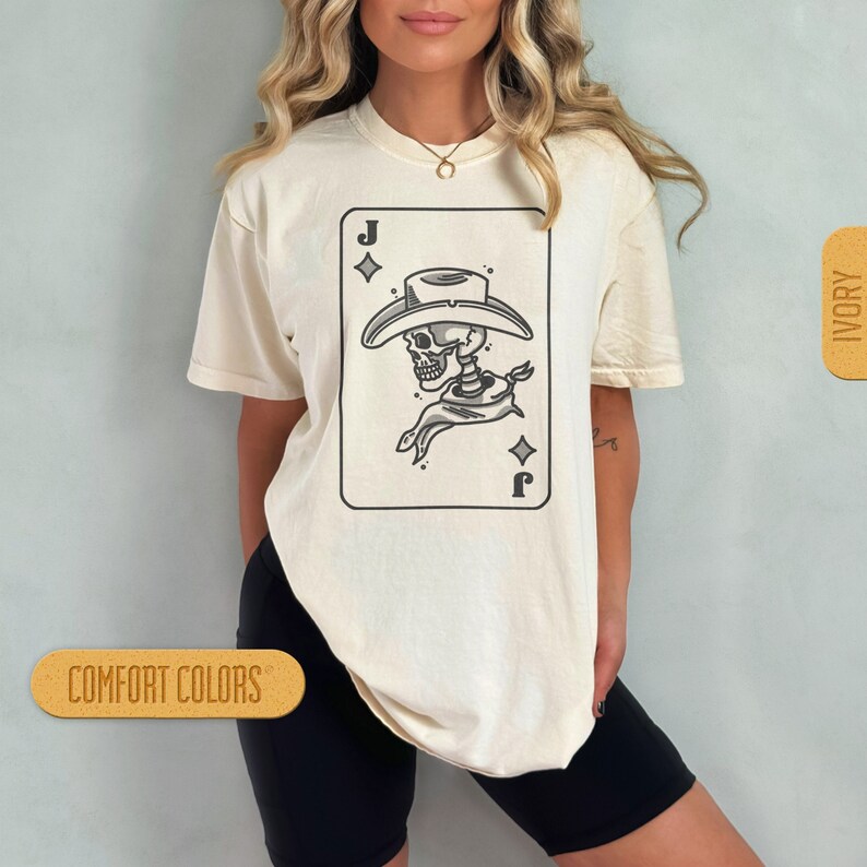 Puede incluir: Una camiseta blanca con un gr&aacute;fico de un esqueleto con un sombrero de vaquero y un pa&ntilde;uelo. El gr&aacute;fico tiene la forma de una carta de juego con la letra 'J' y un s&iacute;mbolo de diamante. La camiseta est&aacute; etiquetada con "COMFORT COLORS" e "IVORY".