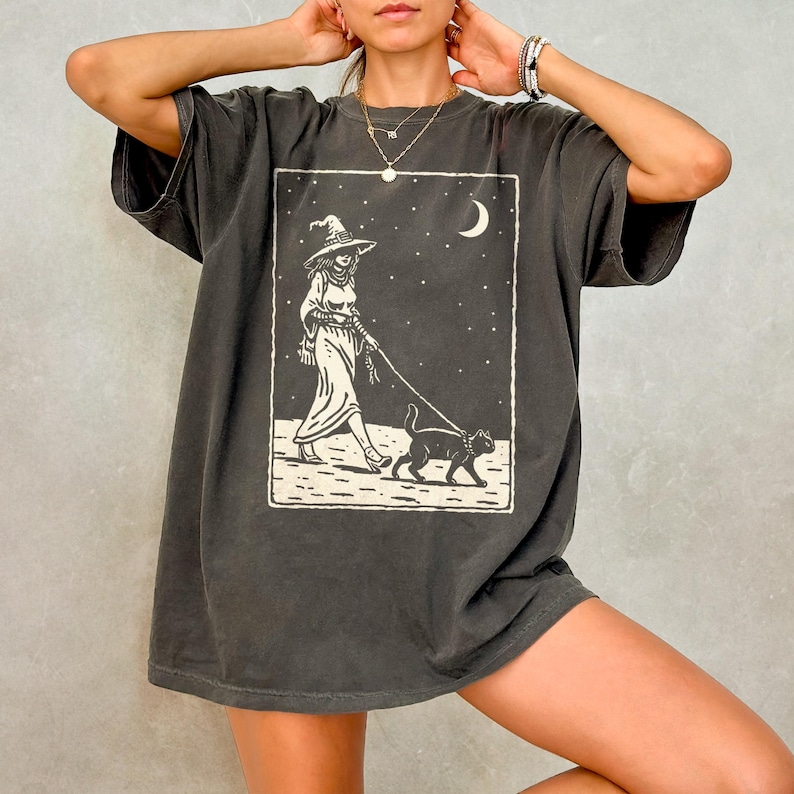 May include: Dark gray t-shirt featuring a witch walking a black cat on a leash under a starry night sky with a crescent moon. The graphic is in a rectangular frame. The shirt has short sleeves and a relaxed fit.