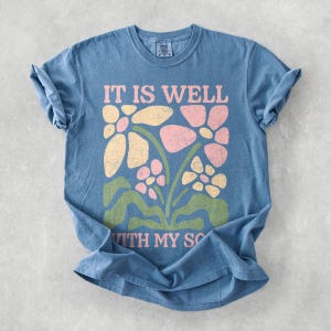 May include: A blue t-shirt with rolled-up sleeves. The shirt features a floral design in pink, yellow, and green, with the text "IT IS WELL WITH MY SO" in pink. The shirt has a vintage, slightly faded appearance.