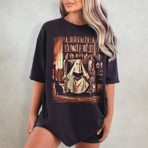 Op de afbeelding: Zwart T-shirt met een afbeelding van een spook dat in een bibliotheek leest. Het spook zit in een fauteuil, met een zwarte kat aan zijn voeten. Een boekenkast en een schedel zijn zichtbaar op de achtergrond. Het shirt heeft een vintage uitstraling.
