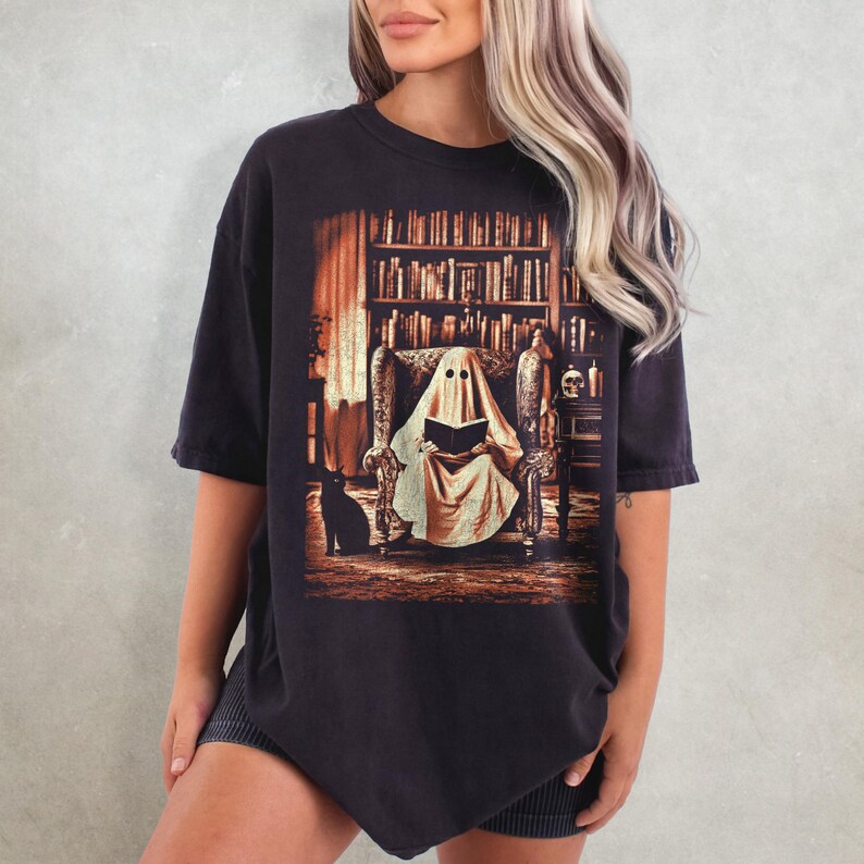 Op de afbeelding: Zwart T-shirt met een afbeelding van een spook dat in een bibliotheek leest. Het spook zit in een fauteuil, met een zwarte kat aan zijn voeten. Een boekenkast en een schedel zijn zichtbaar op de achtergrond. Het shirt heeft een vintage uitstraling.