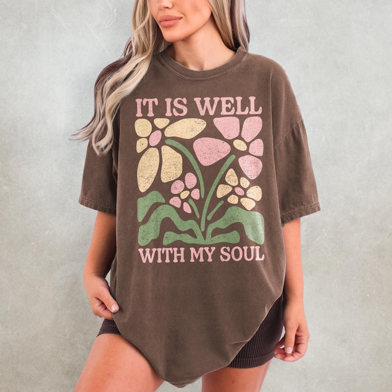 May include: Brown t-shirt with the text "IT IS WELL WITH MY SOUL" in pink. The shirt features a floral design in yellow, pink, and green. The t-shirt has short sleeves and a relaxed fit.