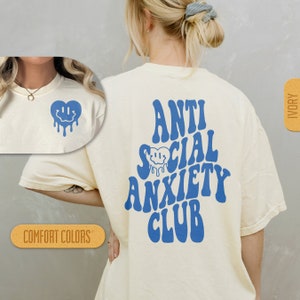 K&ouml;nnte beinhalten: Ein wei&szlig;es T-Shirt mit blauem Text, der "Anti Social Anxiety Club" in einer Retro-Schriftart lautet. Die R&uuml;ckseite des Shirts zeigt ein blaues, tropfendes Smiley-Gesichtsherz.
