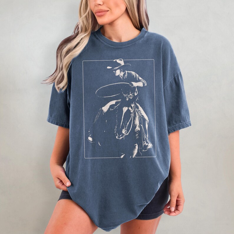 May include: A navy blue oversized t-shirt with a white graphic of a cowboy riding a horse and lassoing a rope.