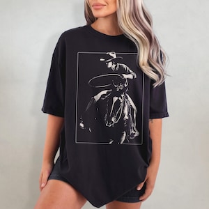 May include: Black oversized t-shirt with a white graphic of a cowboy riding a horse and holding a lasso.