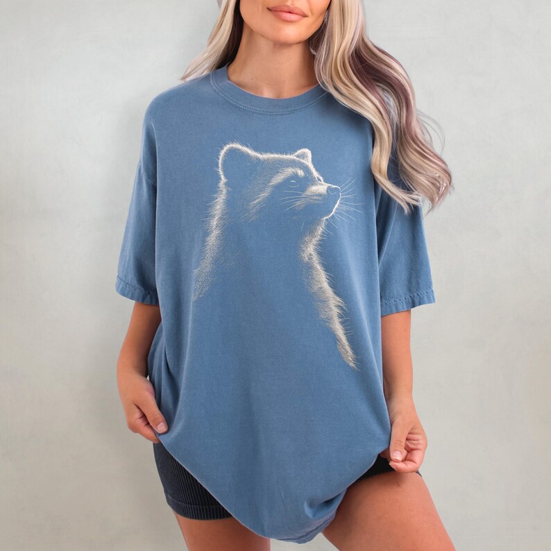 May include: An oversized blue t-shirt featuring a white graphic of a raccoon looking upwards.