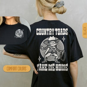 Puede incluir: Camiseta negra con un gr&aacute;fico blanco de una rana de dibujos animados con un sombrero de vaquero y sosteniendo un lazo. El texto "COUNTRY TOADS TAKE ME HOME" est&aacute; impreso en blanco en la camiseta.