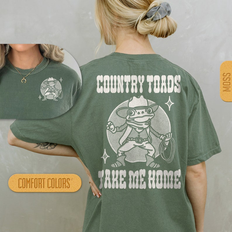 Puede incluir: Una camiseta verde con un gr&aacute;fico blanco de una rana con un sombrero de vaquero y sosteniendo un lazo. El texto "COUNTRY TOADS TAKE ME HOME" est&aacute; impreso en la camiseta.