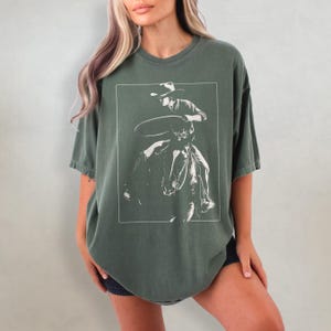 May include: A green oversized t-shirt with a white graphic of a cowboy riding a horse and holding a lasso.