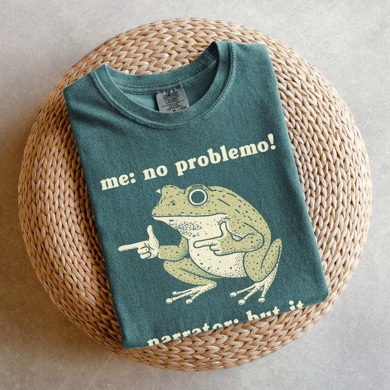 May include: Teal t-shirt featuring a cartoon frog with the text "me: no problemo!" and "narrator: but it". The frog is light green with a white belly and is pointing with its finger. The shirt is folded on a woven, circular surface.