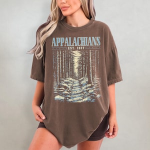 May include: A brown oversized t-shirt with a graphic print of a forest path and the text "Appalachians Est. 1937".