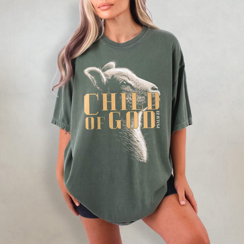 May include: Dark green t-shirt with the text "CHILD OF GOD" in gold lettering and a graphic of a lamb's head. The text "PSALM 23" is printed vertically on the right side. The shirt is a relaxed fit.