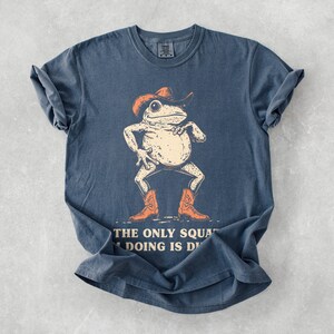 May include: A blue t-shirt featuring a cartoon frog wearing a cowboy hat and boots. The frog is in a confident pose, with the text "THE ONLY SQUAT I'M DOING IS D" below the image. The shirt has a vintage, faded look.