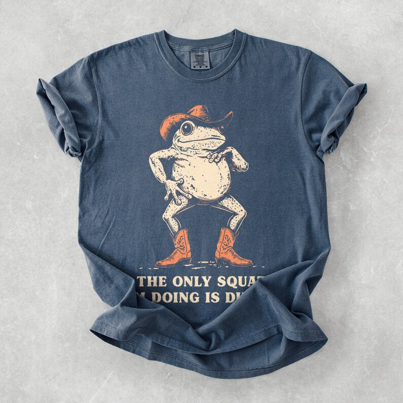 May include: A blue t-shirt featuring a cartoon frog wearing a cowboy hat and boots. The frog is in a confident pose, with the text "THE ONLY SQUAT I'M DOING IS D" below the image. The shirt has a vintage, faded look.