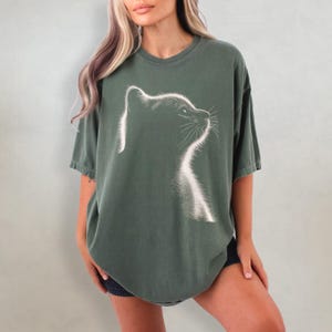 May include: A green oversized t-shirt with a white silhouette of a cat's head looking to the right.
