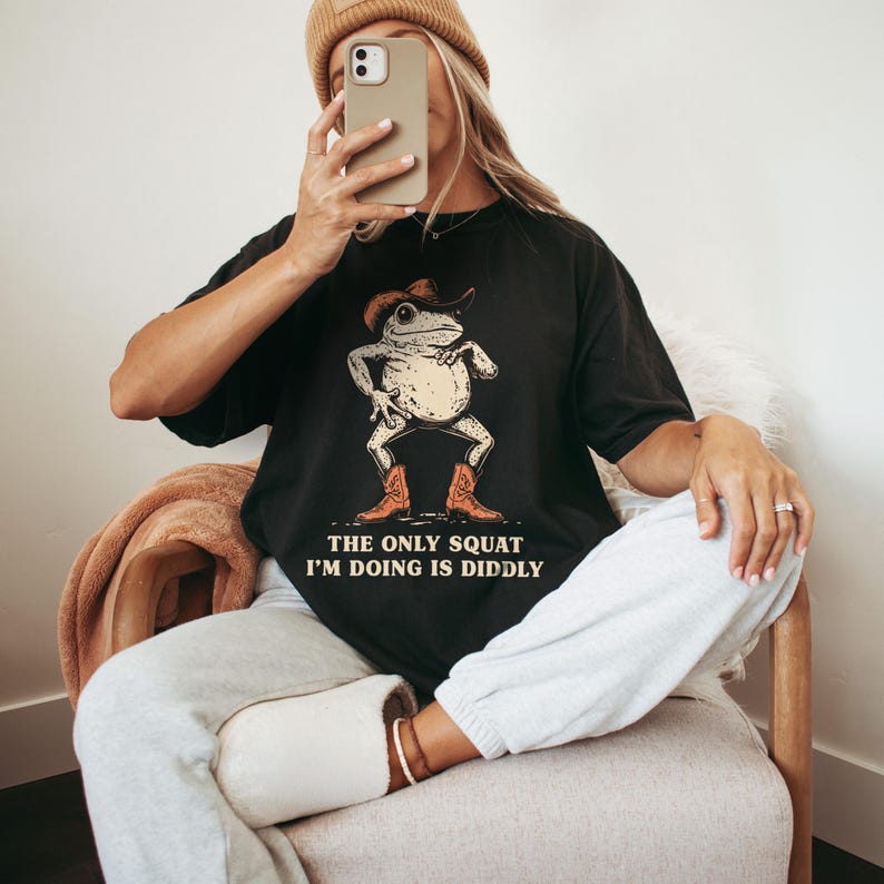 May include: Black t-shirt featuring a cartoon frog wearing a cowboy hat and boots. The frog is in a squatting pose. The text on the shirt reads, "THE ONLY SQUAT I'M DOING IS DIDDLY."