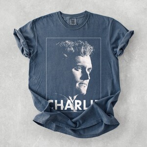 May include: A blue t-shirt with a black and white portrait of a man in a white frame. The name "CHARLI" is printed in white at the bottom. The shirt has rolled sleeves and a crew neck. The fabric appears soft.