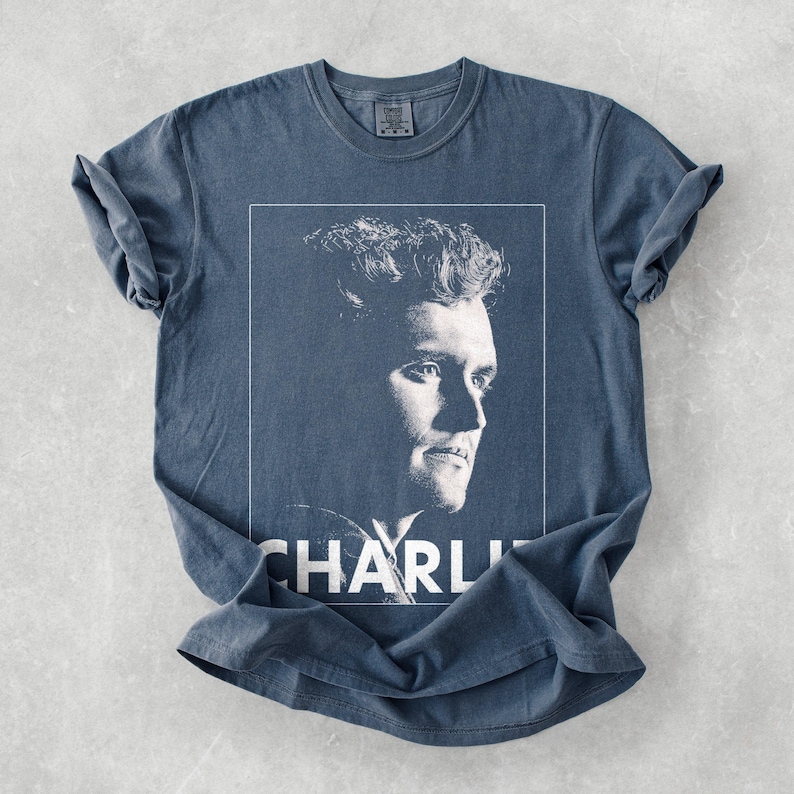 May include: A blue t-shirt with a black and white portrait of a man in a white frame. The name "CHARLI" is printed in white at the bottom. The shirt has rolled sleeves and a crew neck. The fabric appears soft.