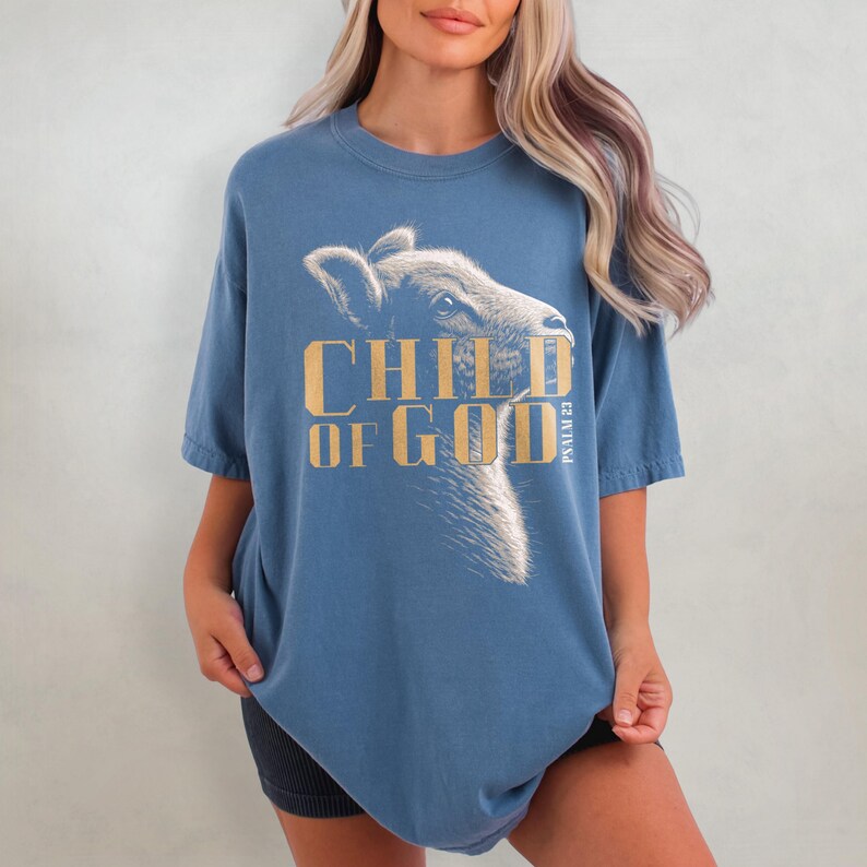 May include: Blue short-sleeved t-shirt with the text "CHILD OF GOD" in gold lettering and a graphic of a calf's head. The text "PSALM 23" is printed vertically on the right side of the graphic.