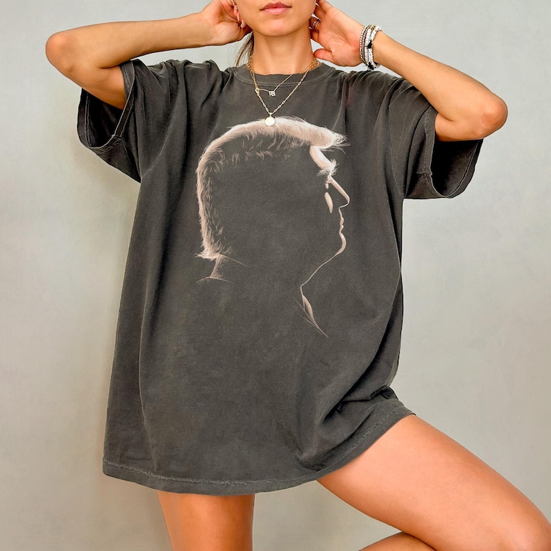 K&ouml;nnte beinhalten: Dunkelgraues, &uuml;bergro&szlig;es T-Shirt mit einem hellen Seitenprofil-Portr&auml;t. Das T-Shirt hat kurze &Auml;rmel und eine lockere Passform. Die helle Grafik kontrastiert mit dem dunklen Stoff, geeignet f&uuml;r Freizeitkleidung.