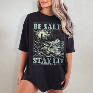 May include: Black oversized t-shirt with a graphic of a lighthouse in a stormy sea. The text "BE SALT STAY LIT" is printed on the shirt, with the scripture reference "MATTHEW 5:13-14" below.