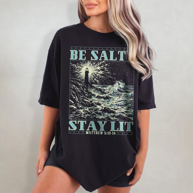 May include: Black oversized t-shirt with a graphic of a lighthouse in a stormy sea. The text "BE SALT STAY LIT" is printed on the shirt, with the scripture reference "MATTHEW 5:13-14" below.