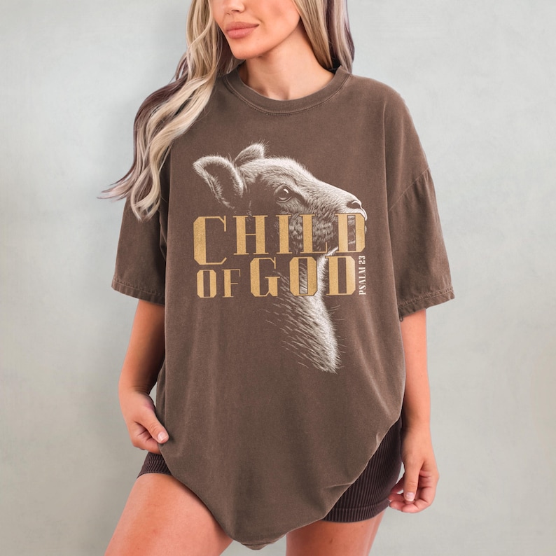 May include: A brown t-shirt with the text "CHILD OF GOD" in gold lettering and a detailed illustration of an animal's head. The shirt also includes the text "PSALM 23".
