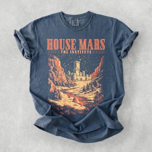 May include: A blue t-shirt with the text "HOUSE MARS THE INSTITUTE" in orange. The graphic depicts a castle scene with red rock formations and a starry night sky. The shirt is made of a soft material.
