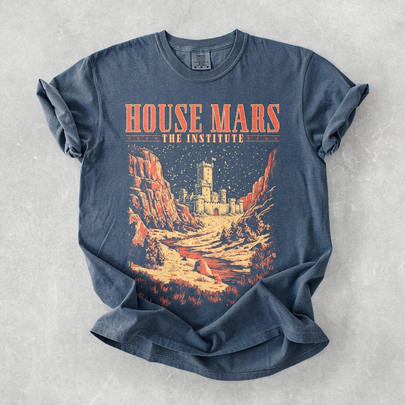 May include: A blue t-shirt with the text "HOUSE MARS THE INSTITUTE" in orange. The graphic depicts a castle scene with red rock formations and a starry night sky. The shirt is made of a soft material.