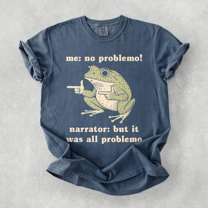 May include: A blue t-shirt with a humorous frog graphic. The frog is green and beige, pointing with both hands. Text on the shirt reads "me: no problemo!" and "narrator: but it was all problemo".