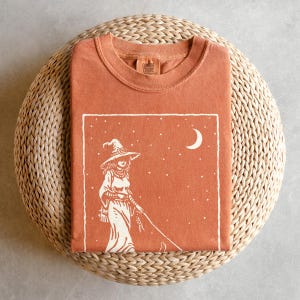 May include: A rust-colored t-shirt with a white graphic of a witch walking with a cat under a crescent moon and stars. The shirt is folded on a woven, circular surface. The brand name "Comfort Colors" is printed on the collar.