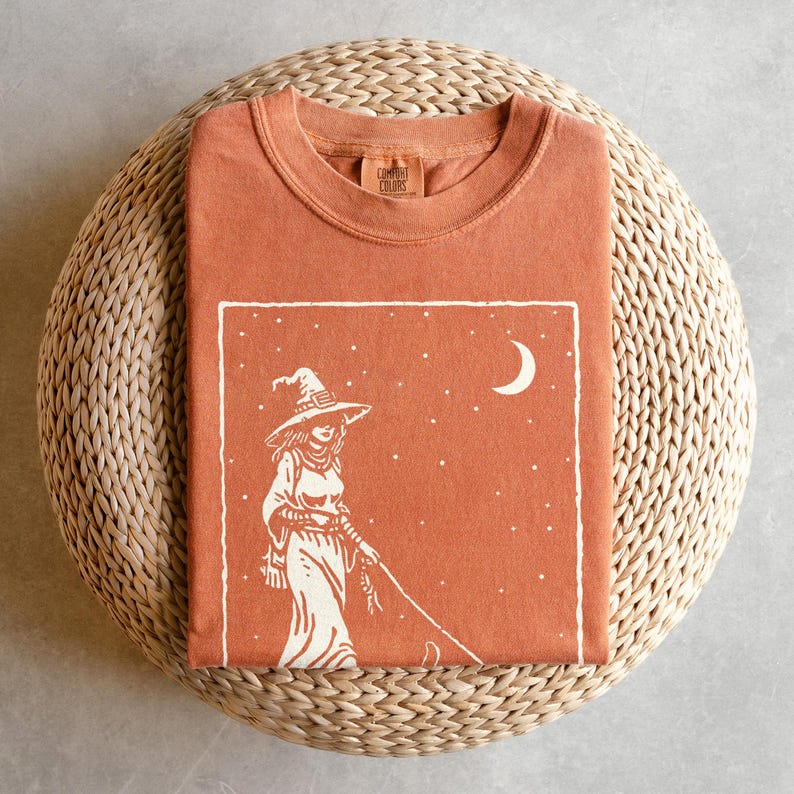May include: A rust-colored t-shirt with a white graphic of a witch walking with a cat under a crescent moon and stars. The shirt is folded on a woven, circular surface. The brand name "Comfort Colors" is printed on the collar.