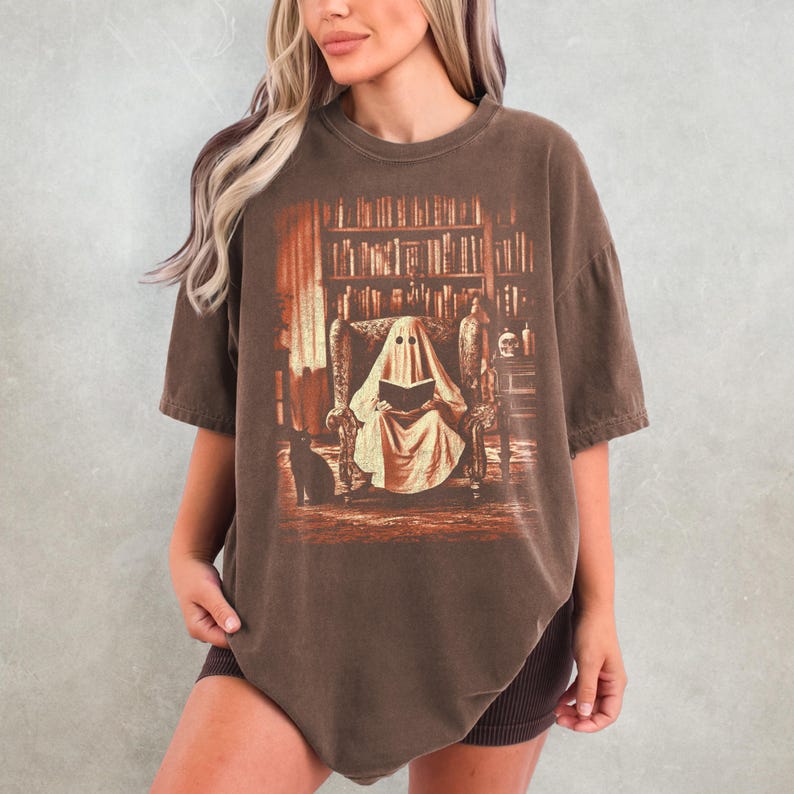 Op de afbeelding: Bruin t-shirt met een grafische print van een geest die een boek leest in een bibliotheek. De geest zit in een fauteuil, met een zwarte kat. De achtergrond toont boekenplanken en antieke decoratie.