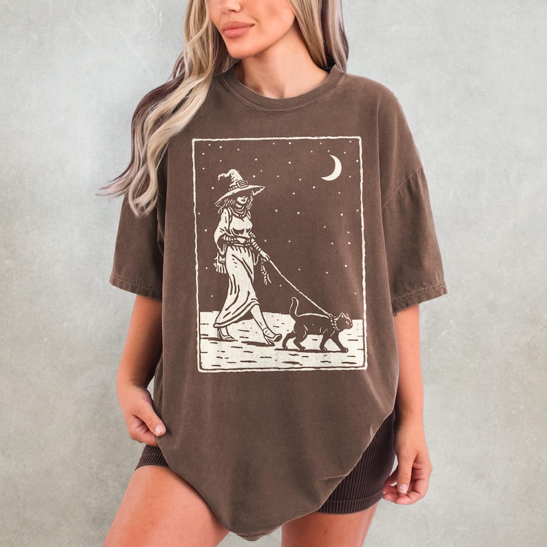 May include: Brown t-shirt featuring a witch walking a cat on a leash under a starry night sky with a crescent moon. The graphic is in a rectangular frame. The witch is wearing a hat and long dress. The shirt is a casual, oversized fit.