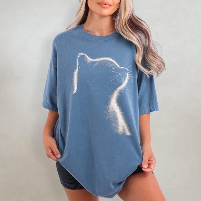 May include: A blue oversized t-shirt with a white outline of a cat's head facing right. The cat's eyes are closed and it has a small, white whisker on the right side of its face.