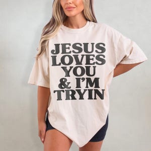 May include: A cream colored t-shirt with the text "JESUS LOVES YOU & I'M TRYIN" printed in black.